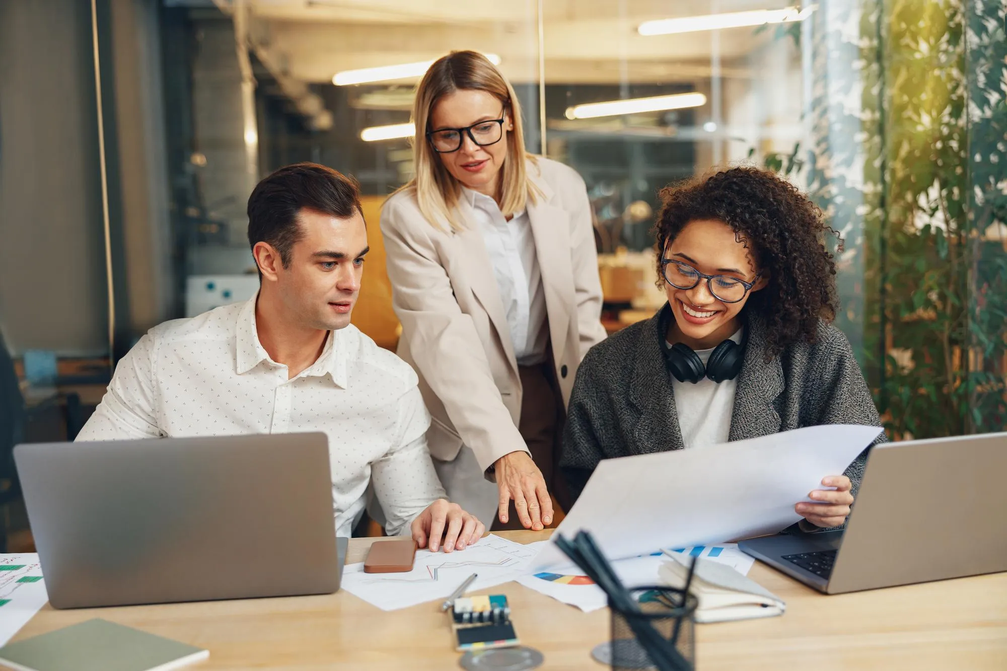 Three diverse professionals collaborating in a modern office, reviewing printed data charts and graphs on a desk with laptops.