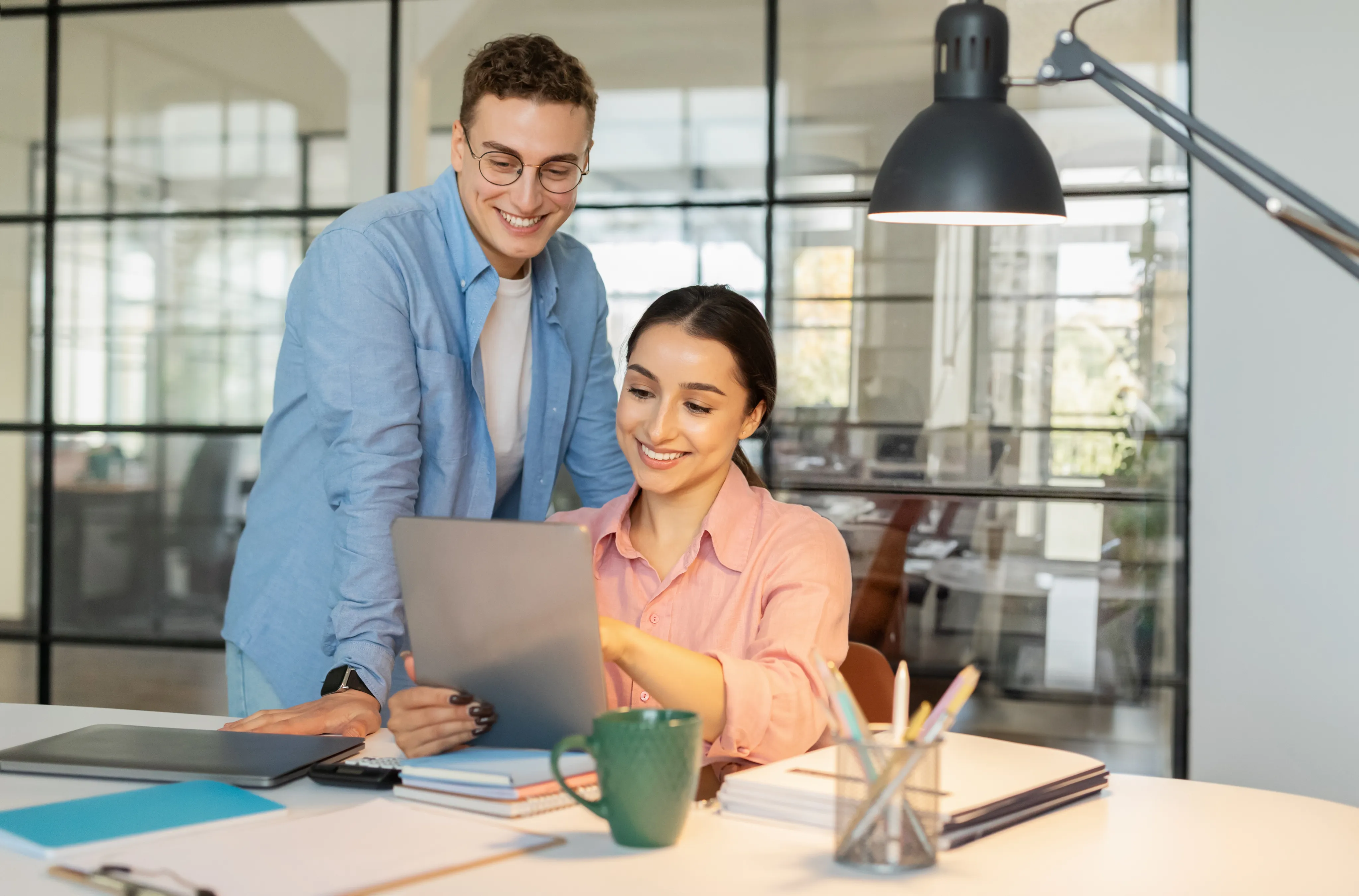 Two smiling coworkers collaborating on a digital tablet at a desk in a bright, modern office with large glass windows.
