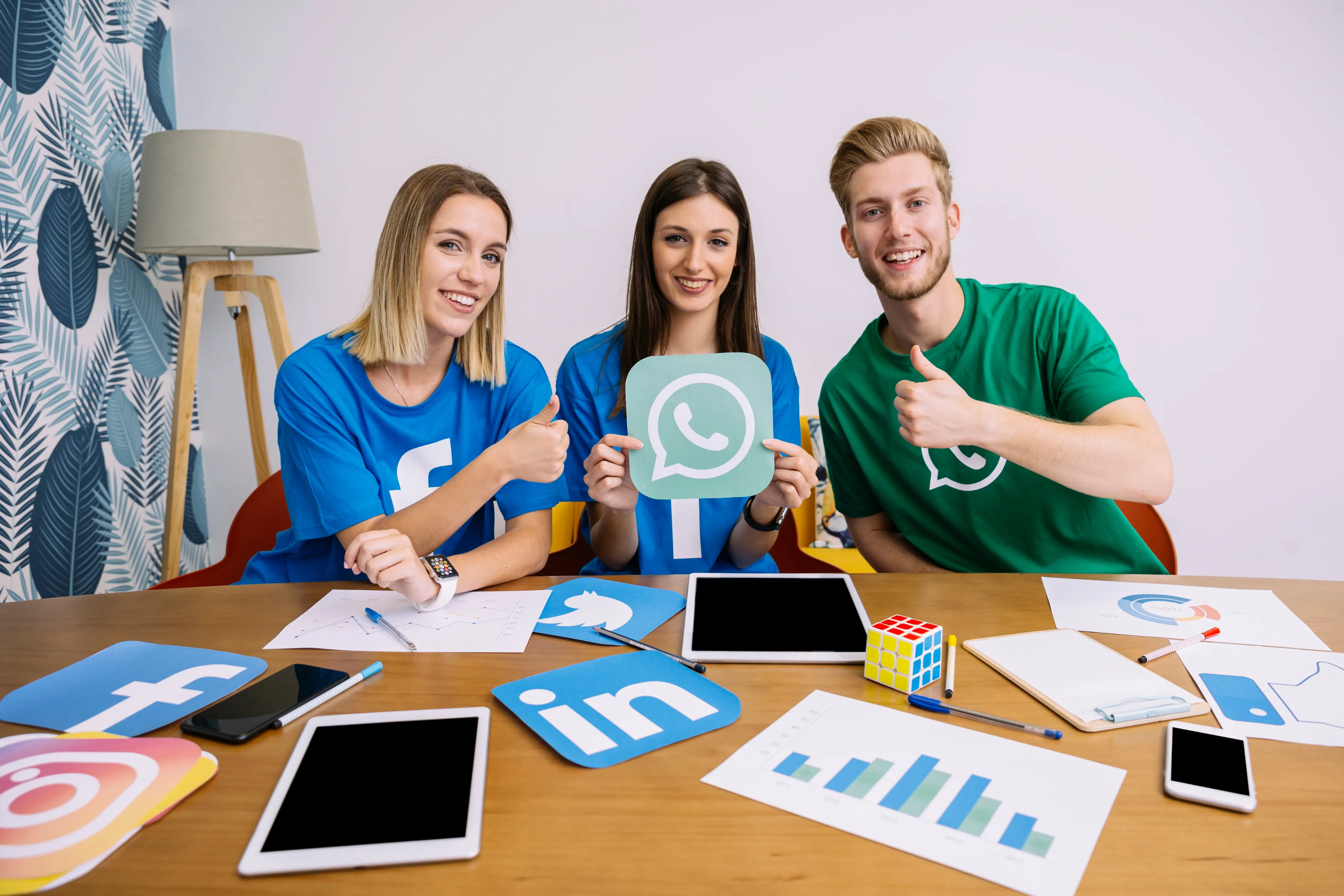 Lilac FloweThree young smiling social media marketing professionals sitting at a desk covered in analytics reports, tablets, and social media logos for Facebook, LinkedIn, Instagram, and WhatsApp.r