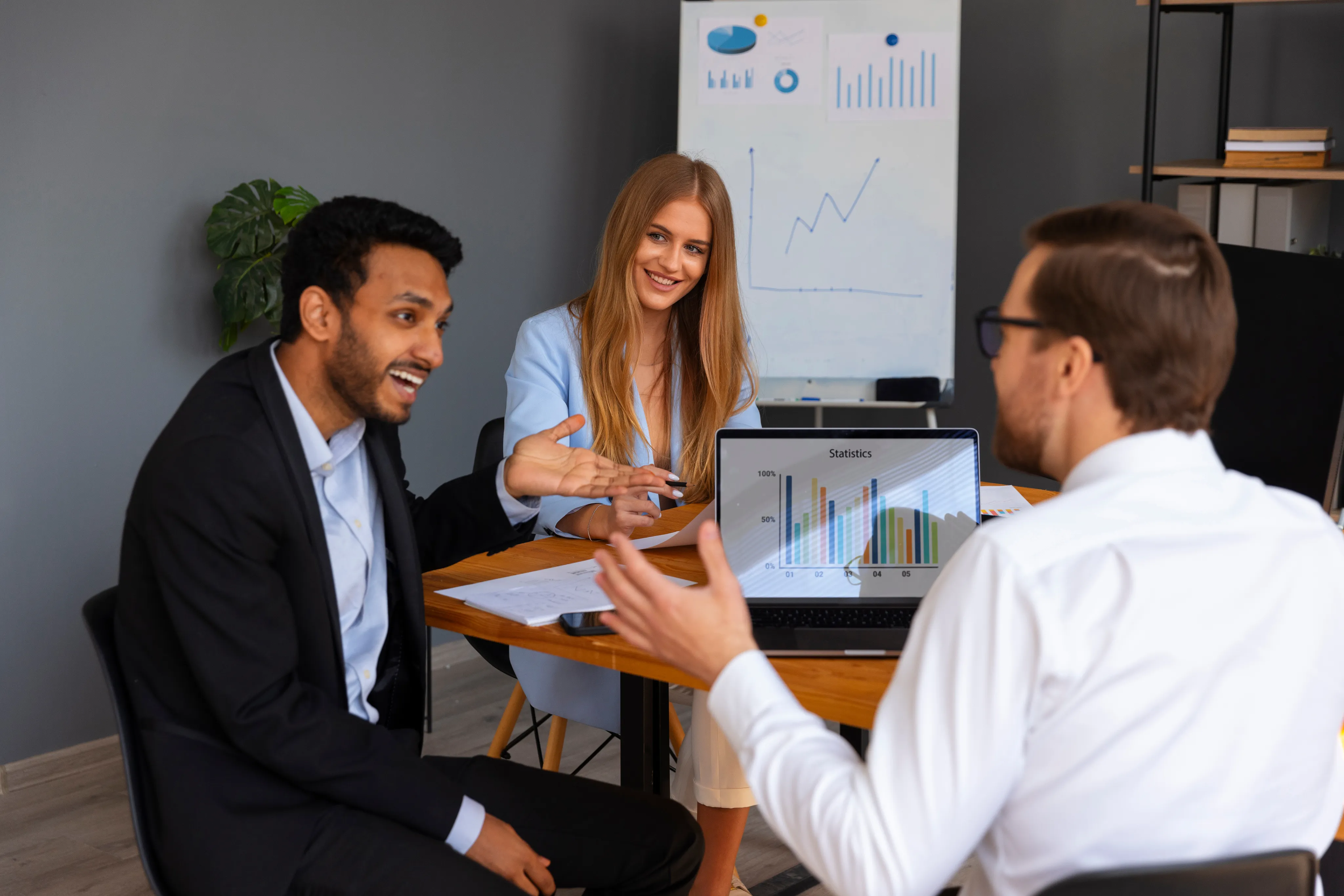 Three colleagues in a meeting room discuss data. A laptop shows colorful graphs, and a whiteboard behind them displays charts. The mood is collaborative.