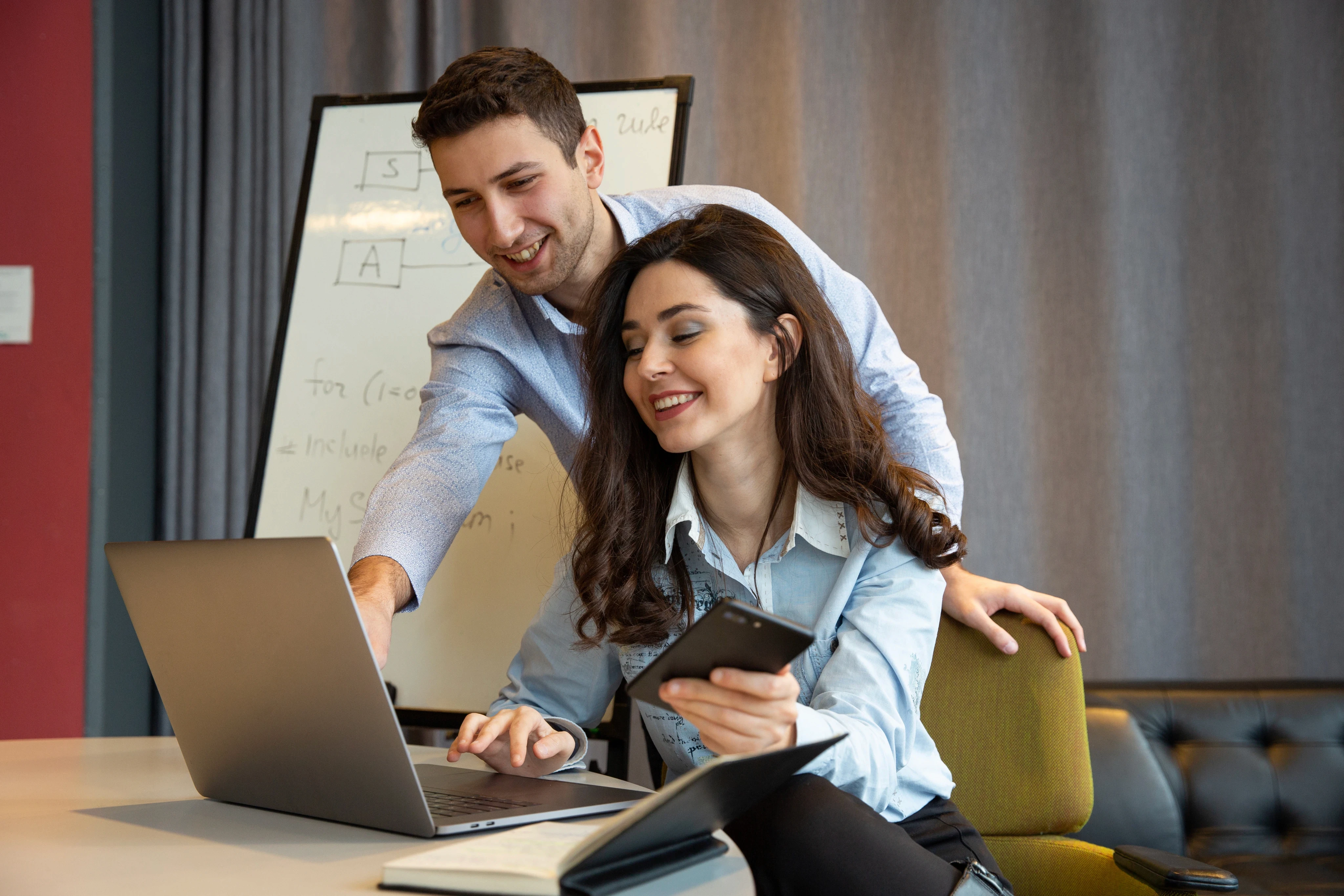 Two smiling colleagues collaborating on a laptop in a modern office with a flowchart on a whiteboard in the background.