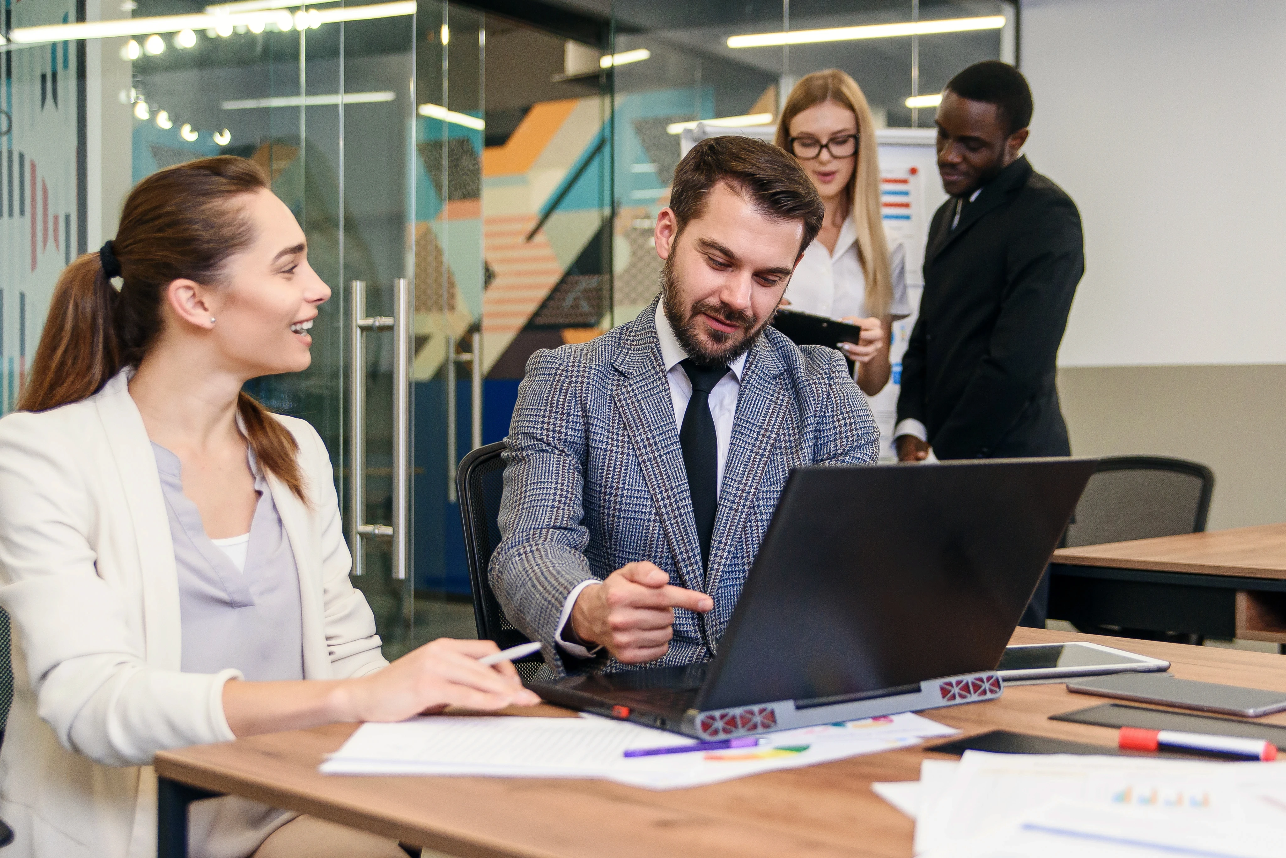 Two people sit at a desk with a laptop, engaged in a discussion, in a modern office. Two others converse in the background, creating a collaborative atmosphere.