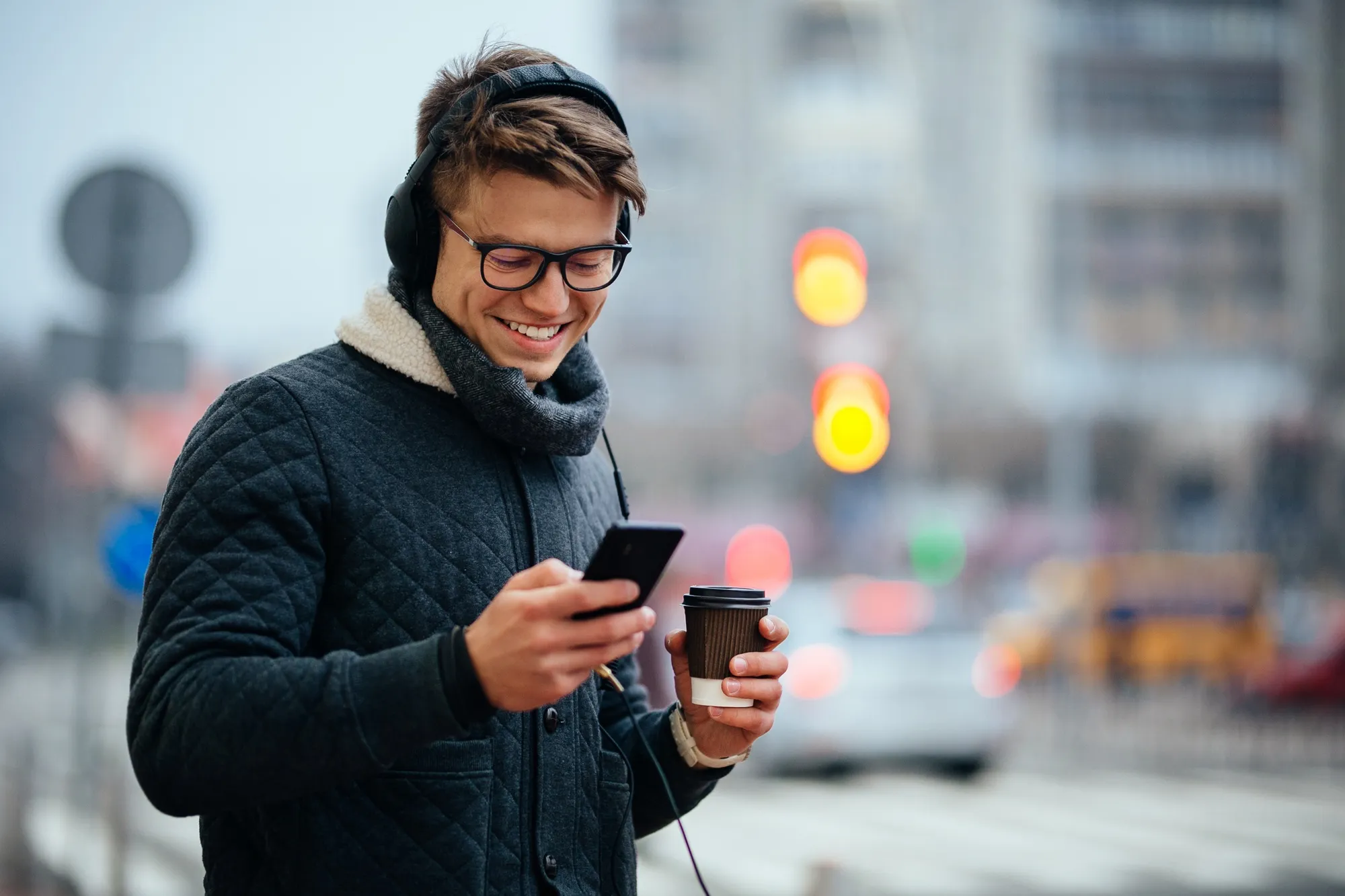 A smiling young man wearing headphones and glasses using a smartphone while holding a coffee cup on a blurred city street.