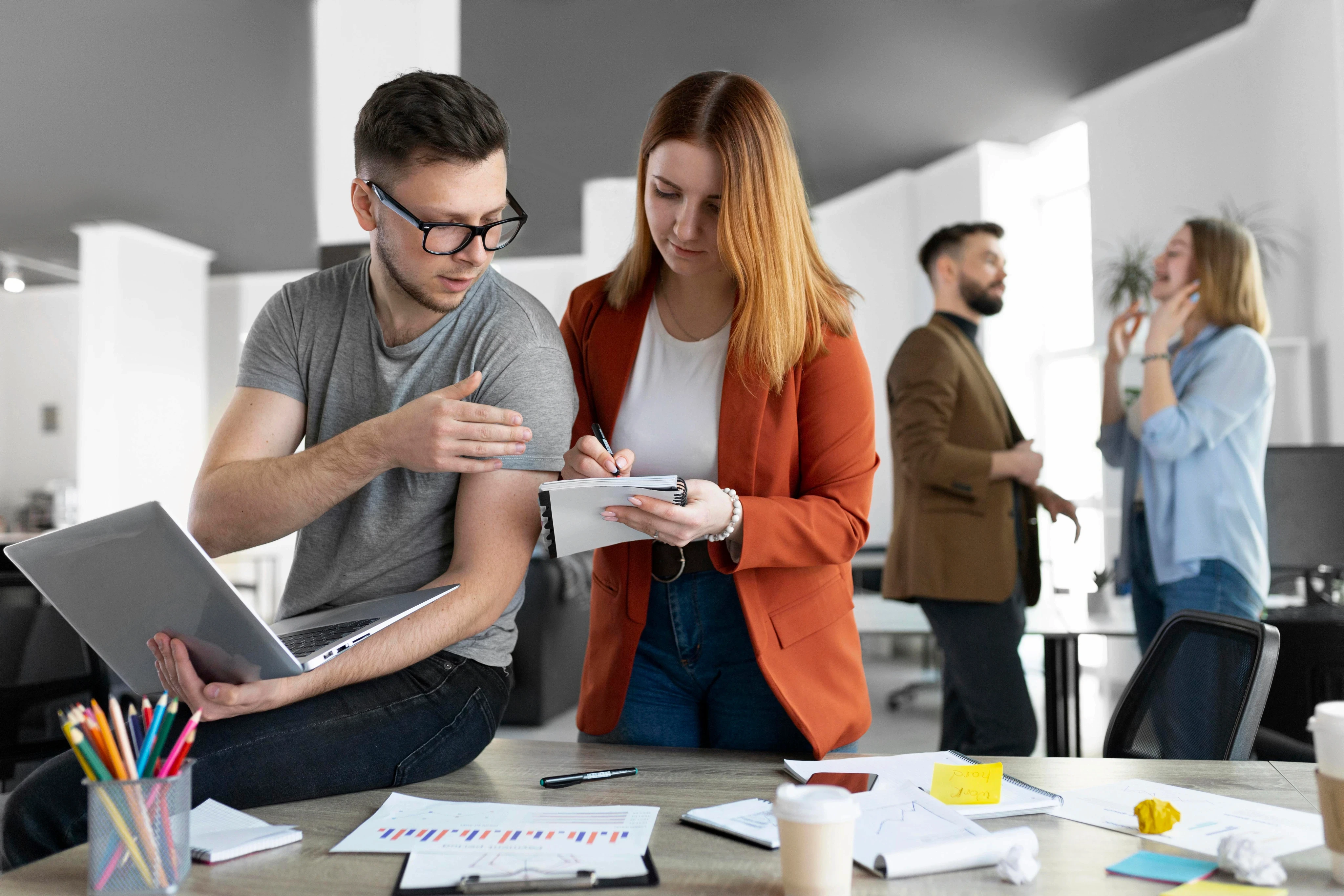 Two young business professionals collaborating in a bright, open-plan office, with one person holding a laptop while the other takes notes in a notebook.