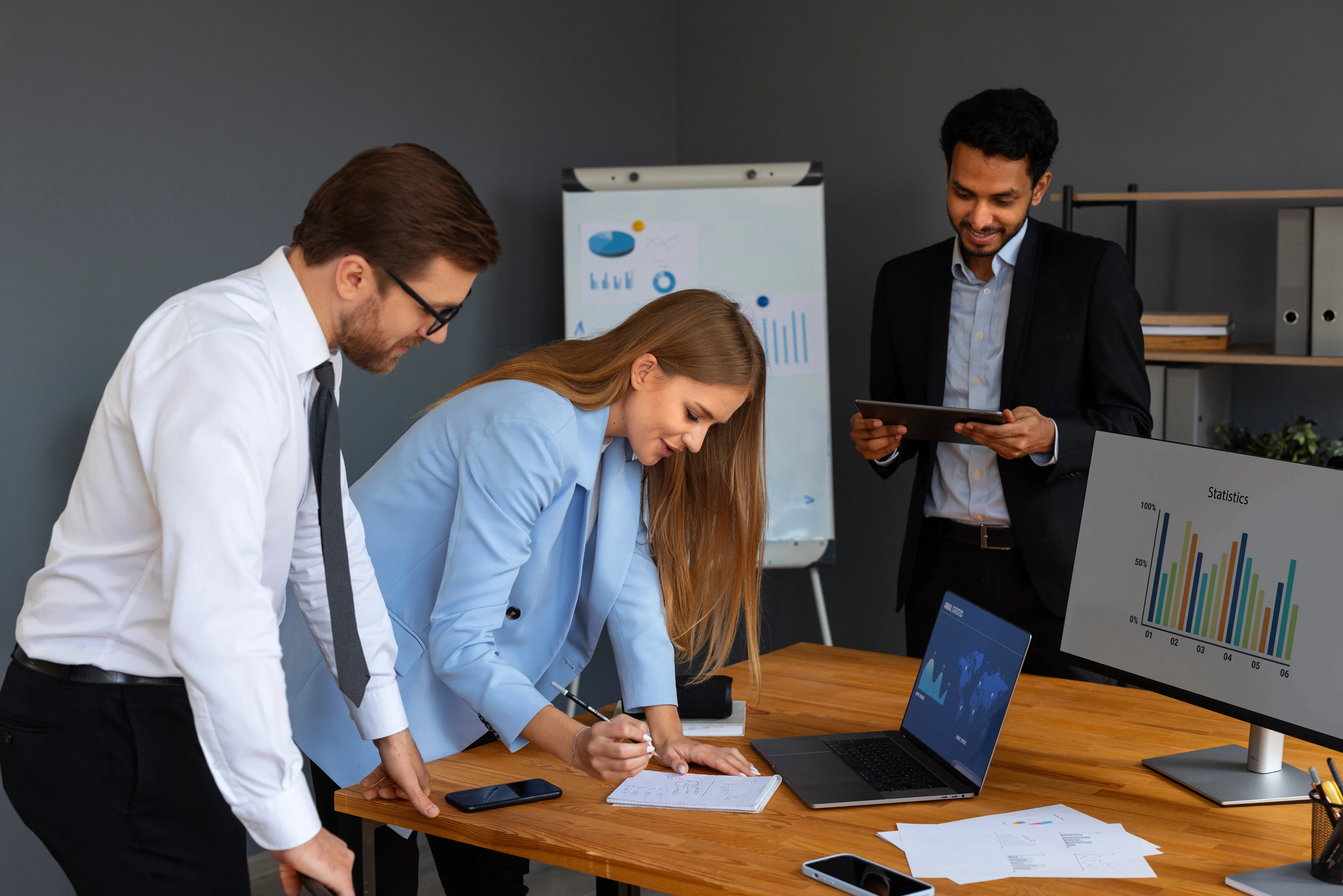 Three young business professionals collaborating in a modern office, analyzing data on a computer monitor and laptop while taking notes.