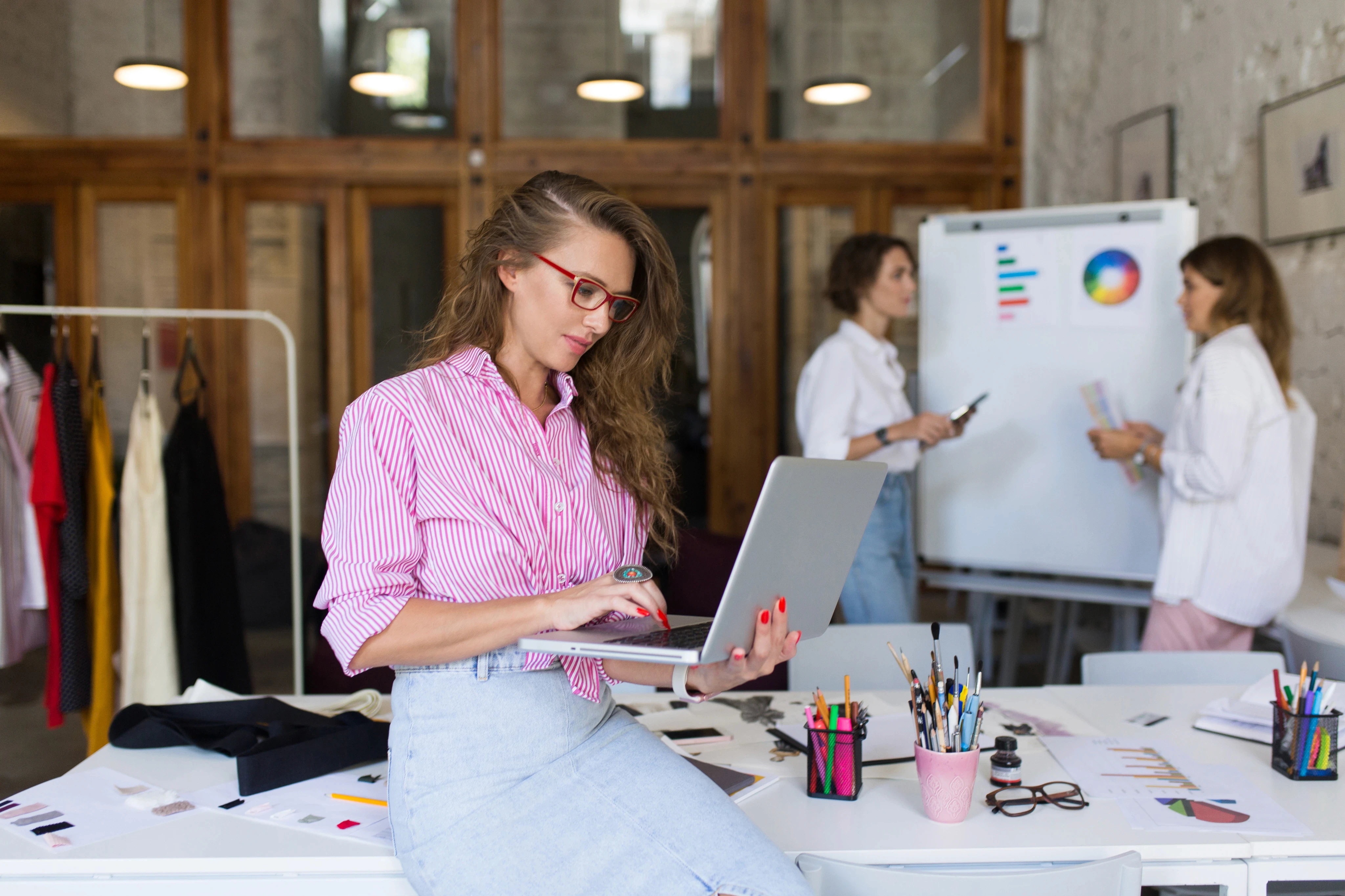 A creative fashion designer in a pink striped shirt working on a laptop while sitting on a desk in a studio, with colleagues reviewing charts on a whiteboard in the background.