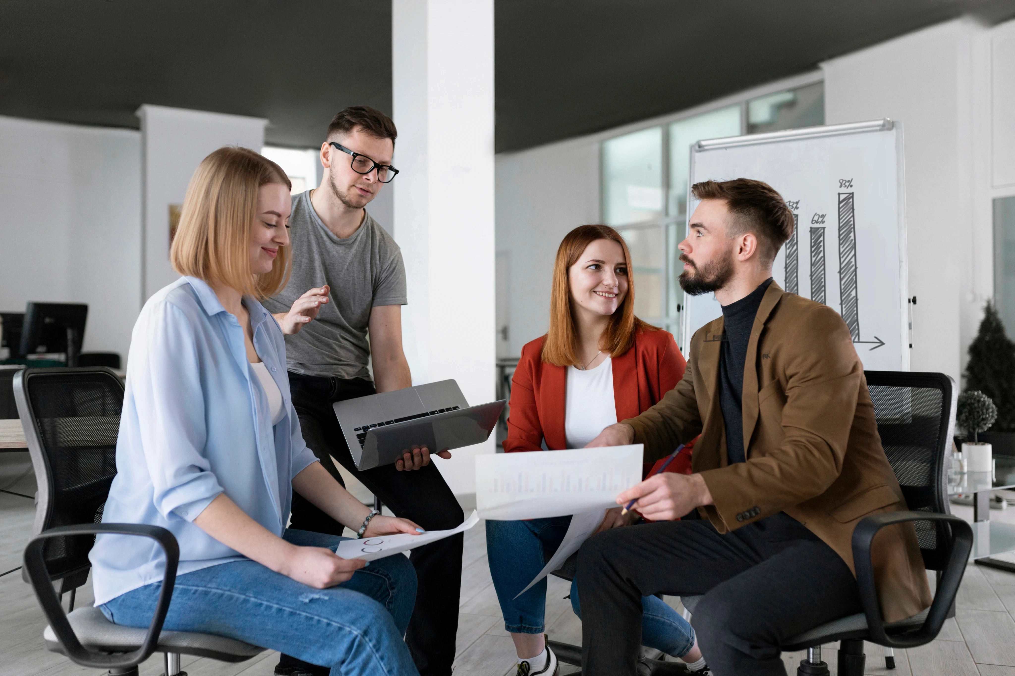 A diverse team of young professionals holding a business meeting in a modern open-plan office, reviewing data reports and a laptop.