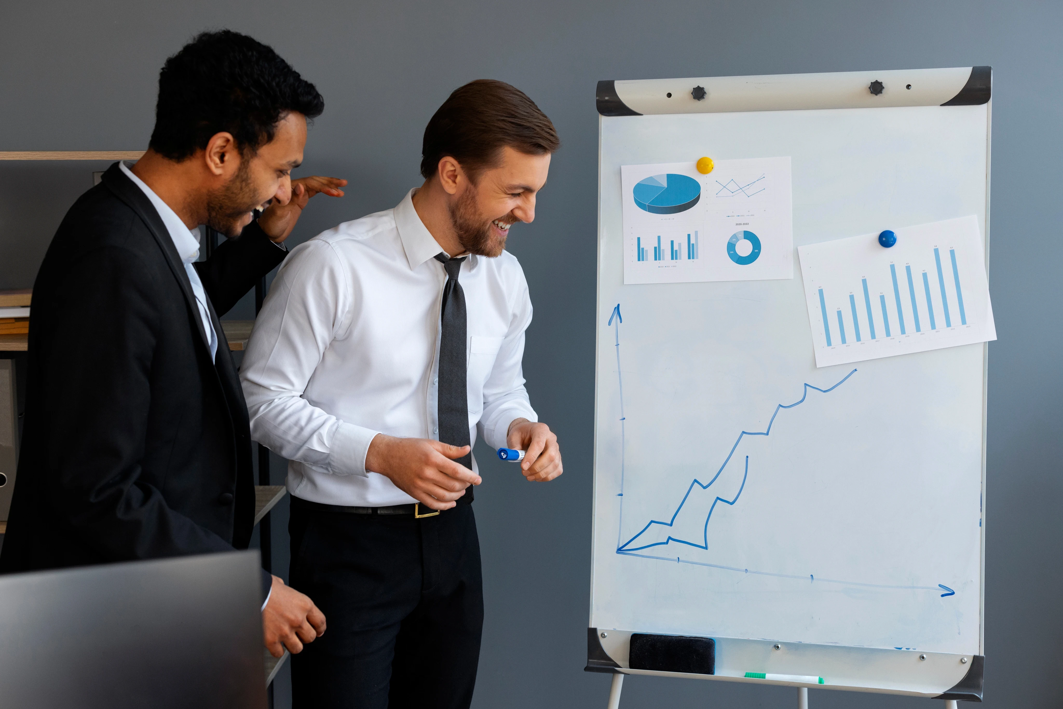Two businessmen celebrating and laughing in front of a whiteboard displaying a rising growth chart and data infographics in an office.
