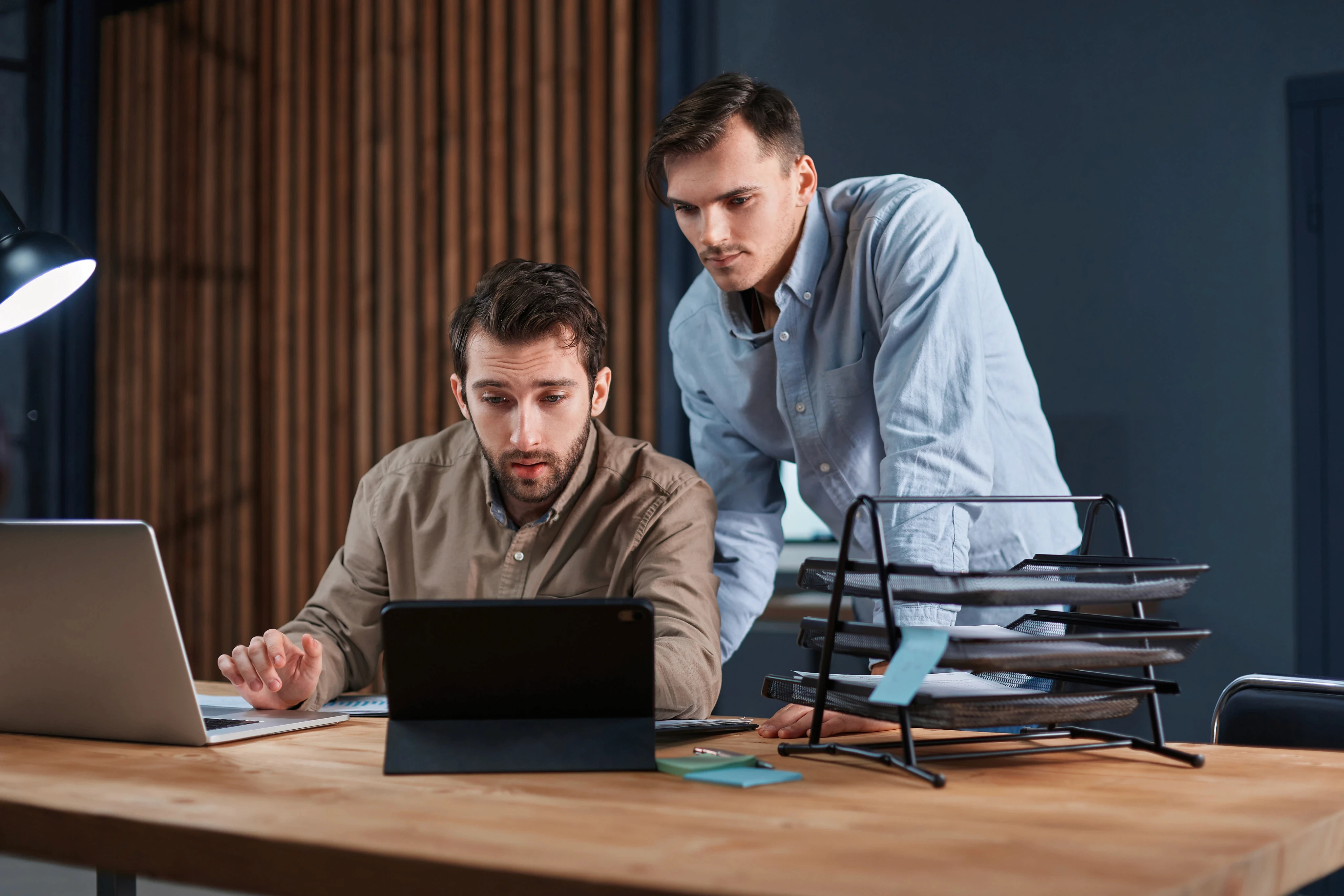 Two focused male colleagues collaborating at a wooden desk in a modern office, looking closely at a tablet and laptop screen while working together.