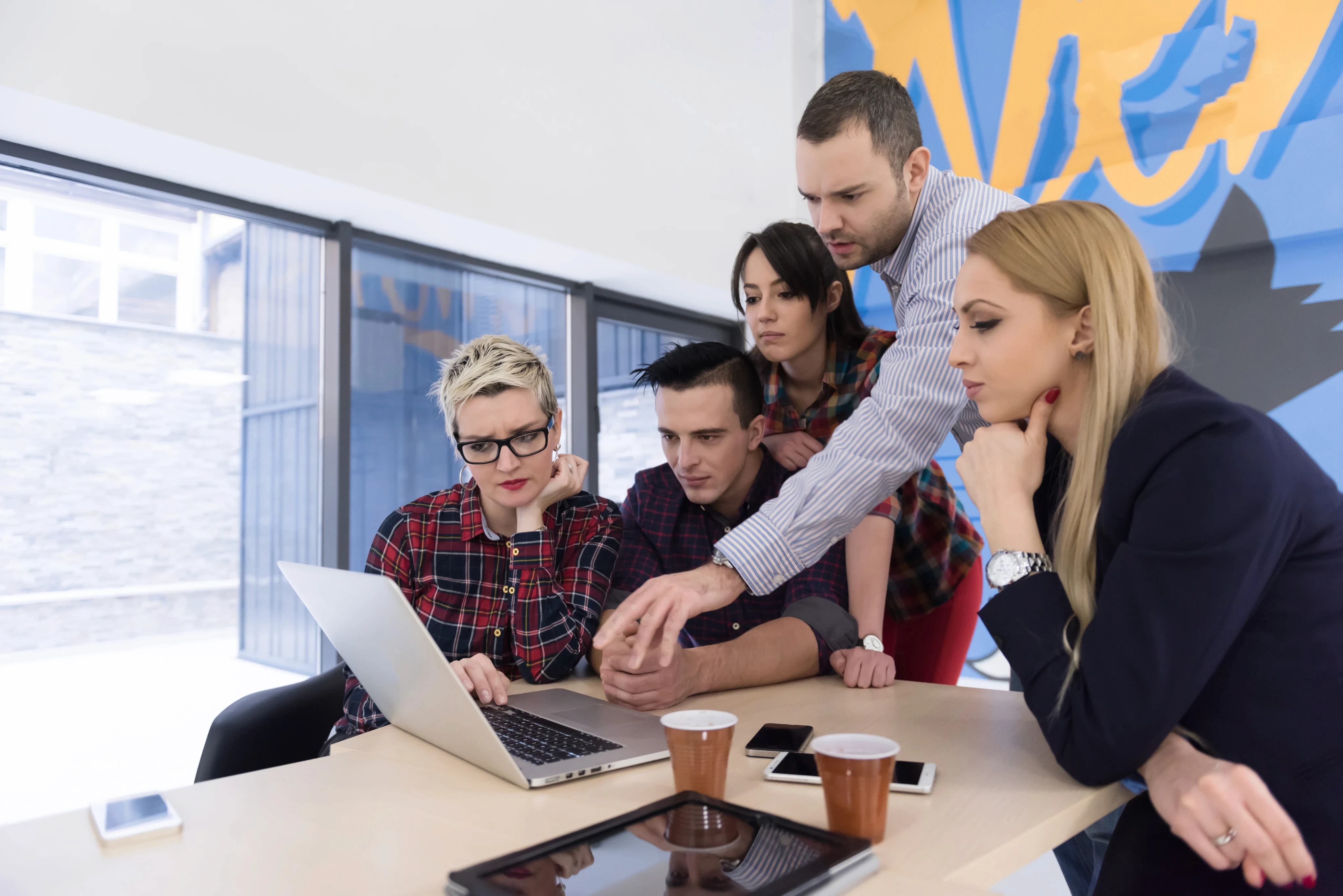 A group of focused creative professionals collaborating around a laptop in a bright, modern office space.
